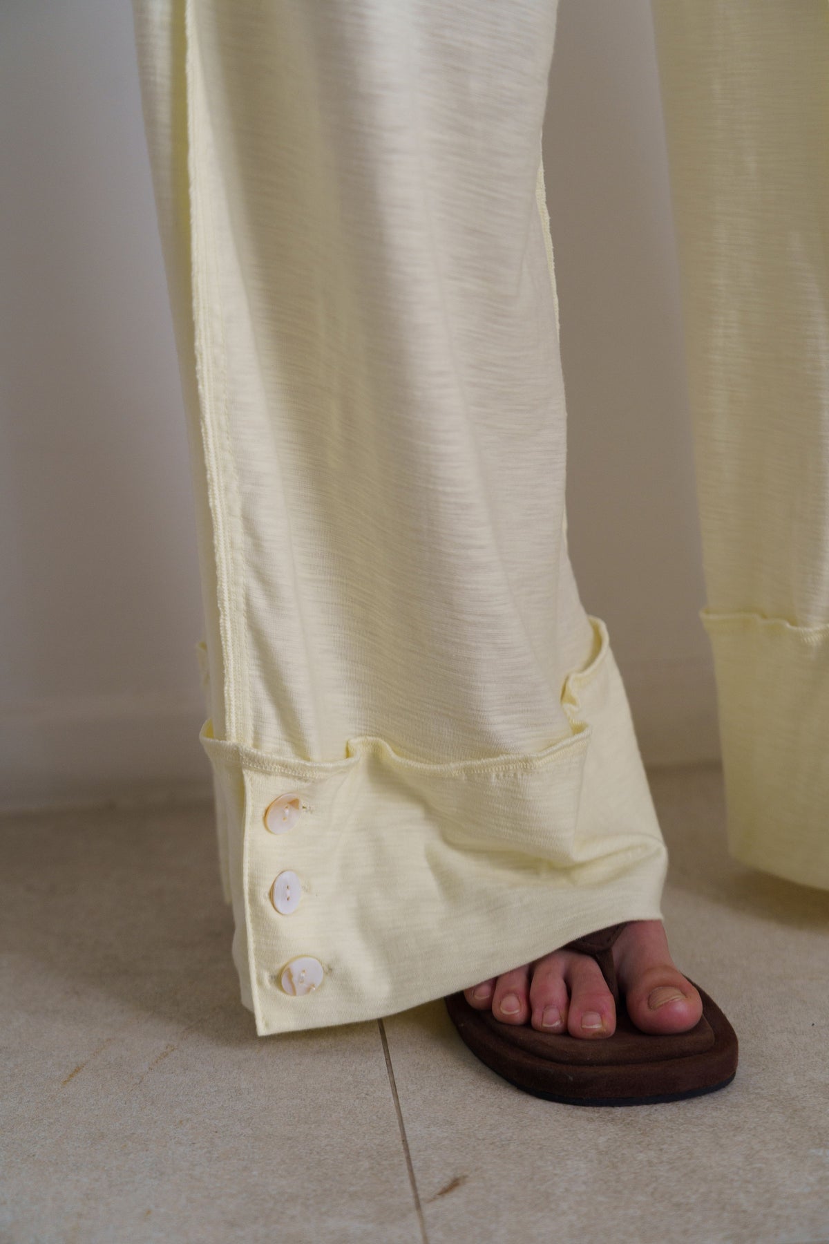 Close-up of a person wearing pale yellow wide-leg trousers with rolled cuffs and decorative buttons, paired with a dark brown flip-flop, standing on a light tiled floor.