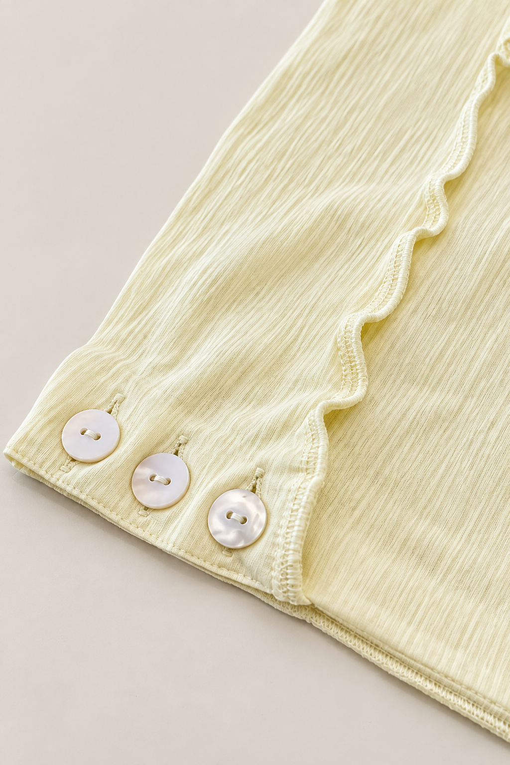 Close-up of a pale yellow textured fabric garment with a scalloped ruffle trim and three round white buttons on the cuff, set against a light beige background.