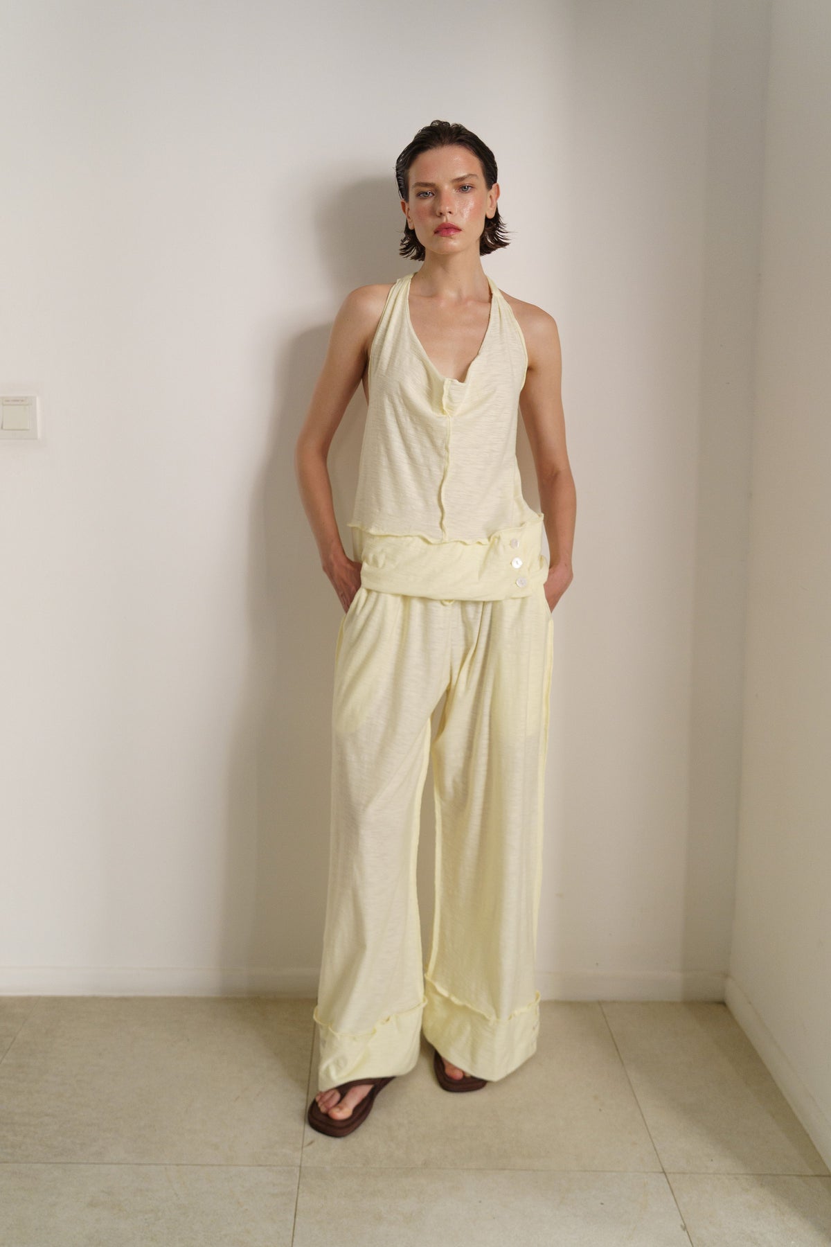 A model stands in a minimalist room with off-white walls and tiled floors, wearing a pale yellow sleeveless halter top and matching wide-leg pants.