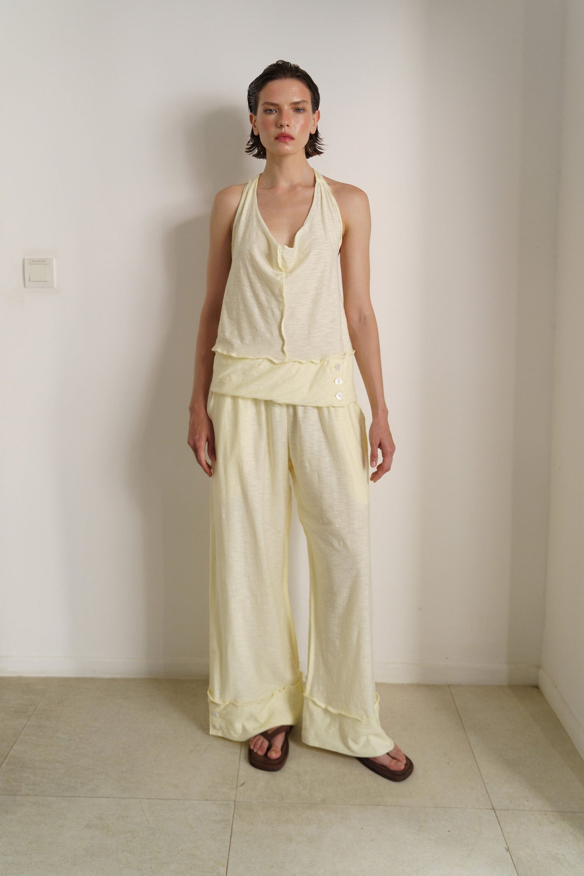 A model stands in a minimalist room wearing a pale yellow linen two-piece set consisting of a halter-neck top and matching wide-leg pants.