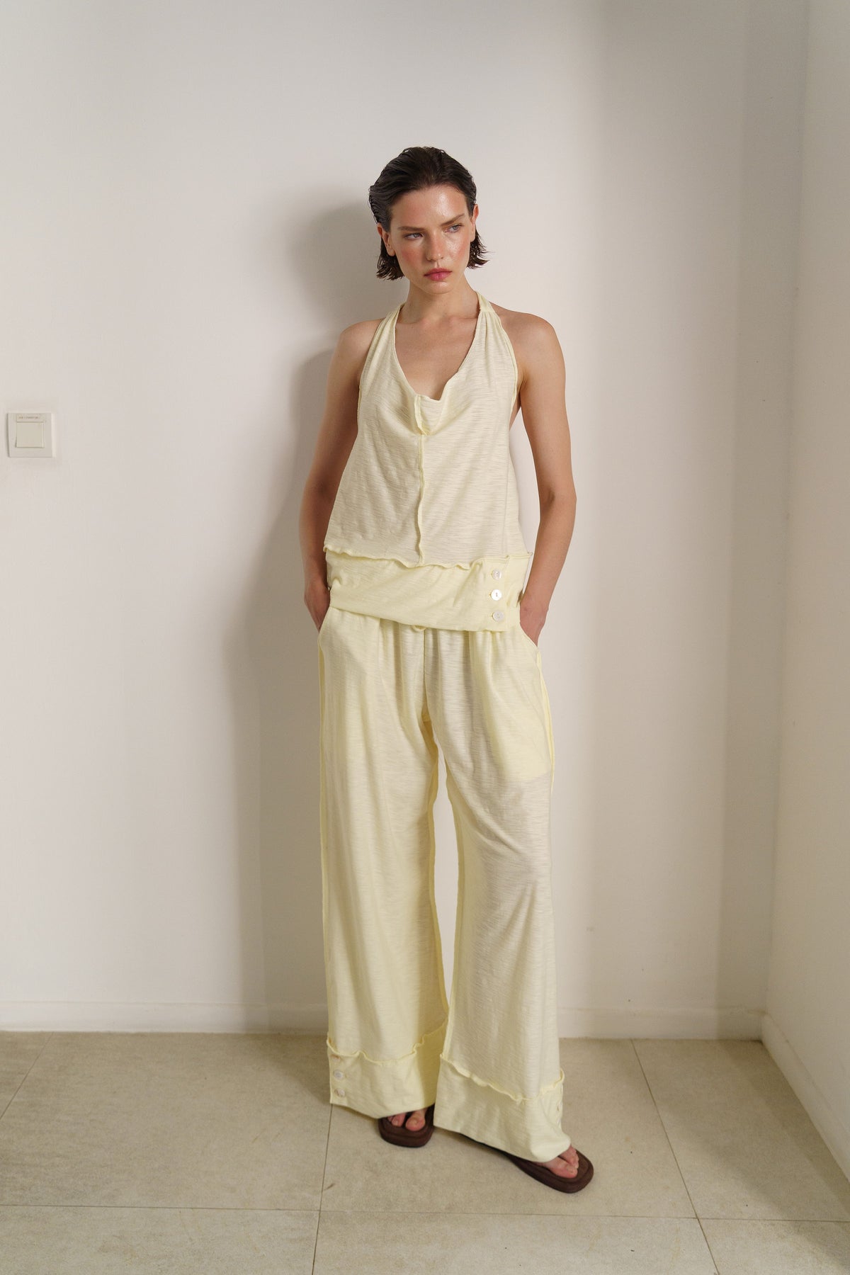 A model stands in a minimalist room wearing a pale yellow sleeveless halter top and matching wide-leg pants.