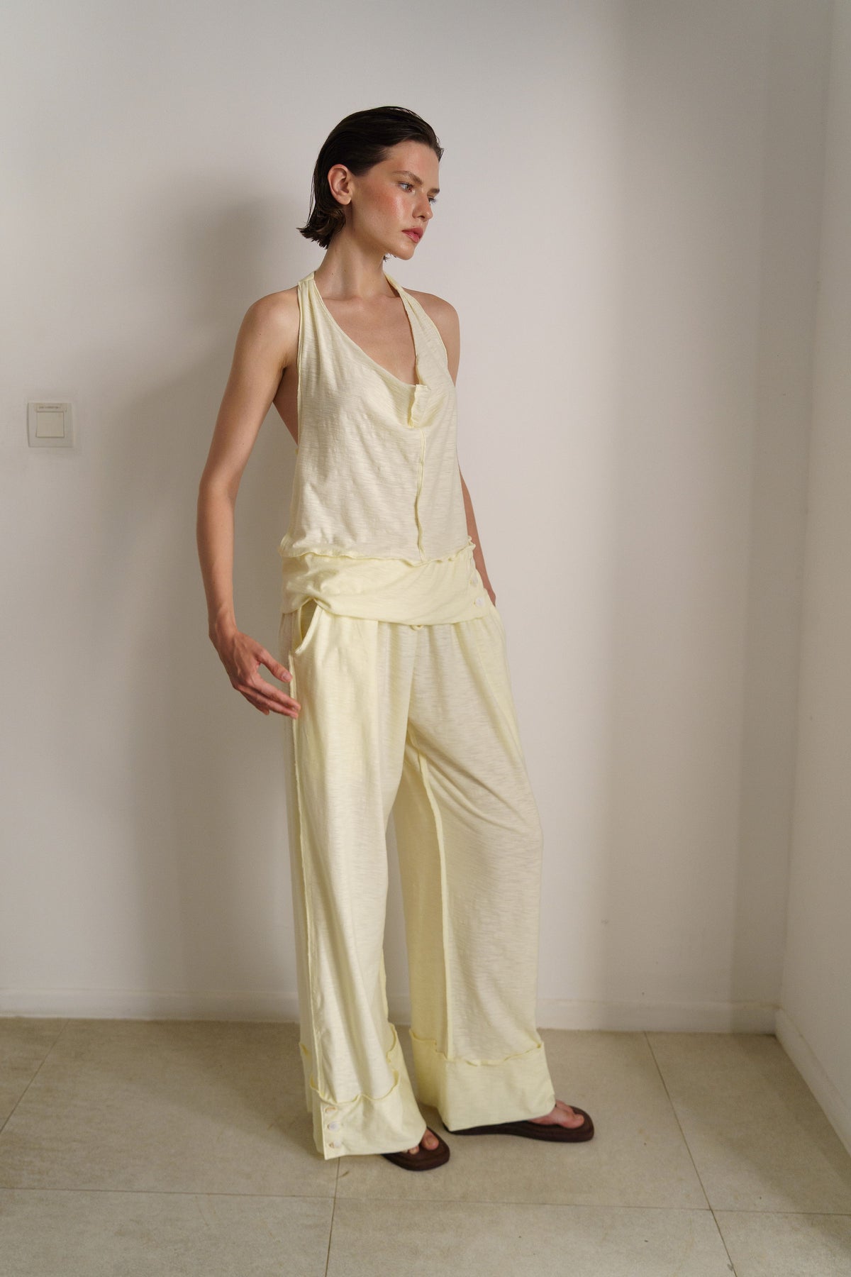 A woman modeling a pale cream two-piece linen set consisting of a halter top and matching wide-leg pants, standing in a minimalist room with off-white walls and light beige tiled floors.