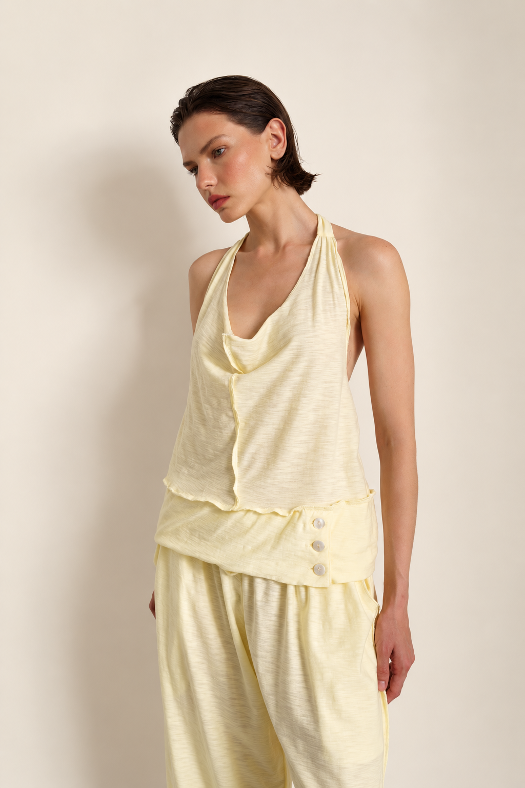 A model wearing a pale yellow two-piece loungewear set consisting of a halter-neck top with a ruffled front and matching wide-leg pants, set against a plain beige studio background.