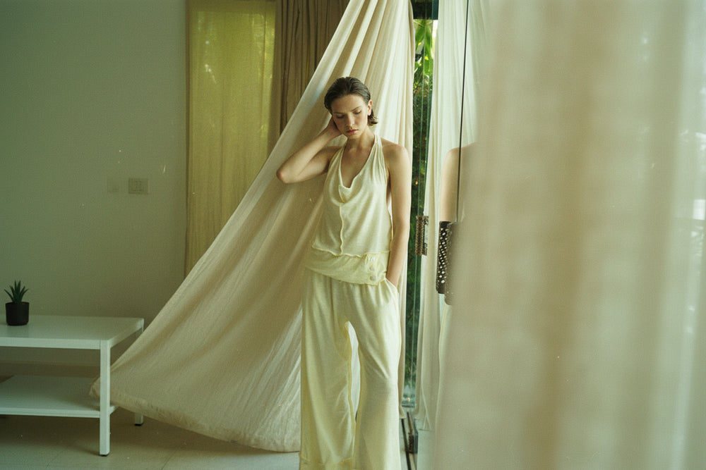 A woman stands in a softly lit room wearing a light cream satin two-piece set consisting of a sleeveless top and wide-leg pants. A large cream fabric drape hangs from a window behind her, and a mirror on the right reflects part of the interior.