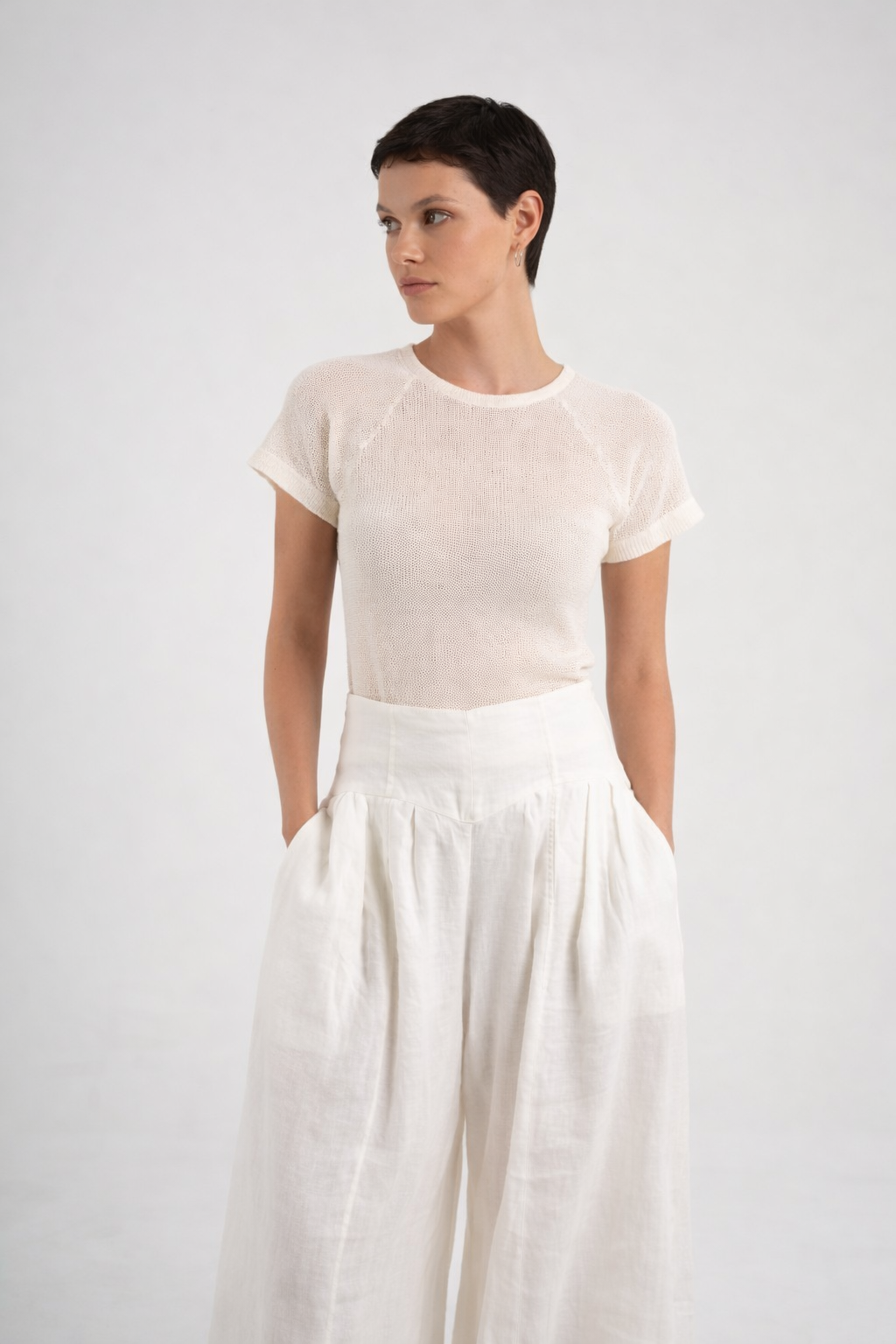 A model wearing a short-sleeve crewneck top in a semi-sheer off-white fabric paired with high-waisted, wide-leg white linen trousers, set against a plain light gray studio background.