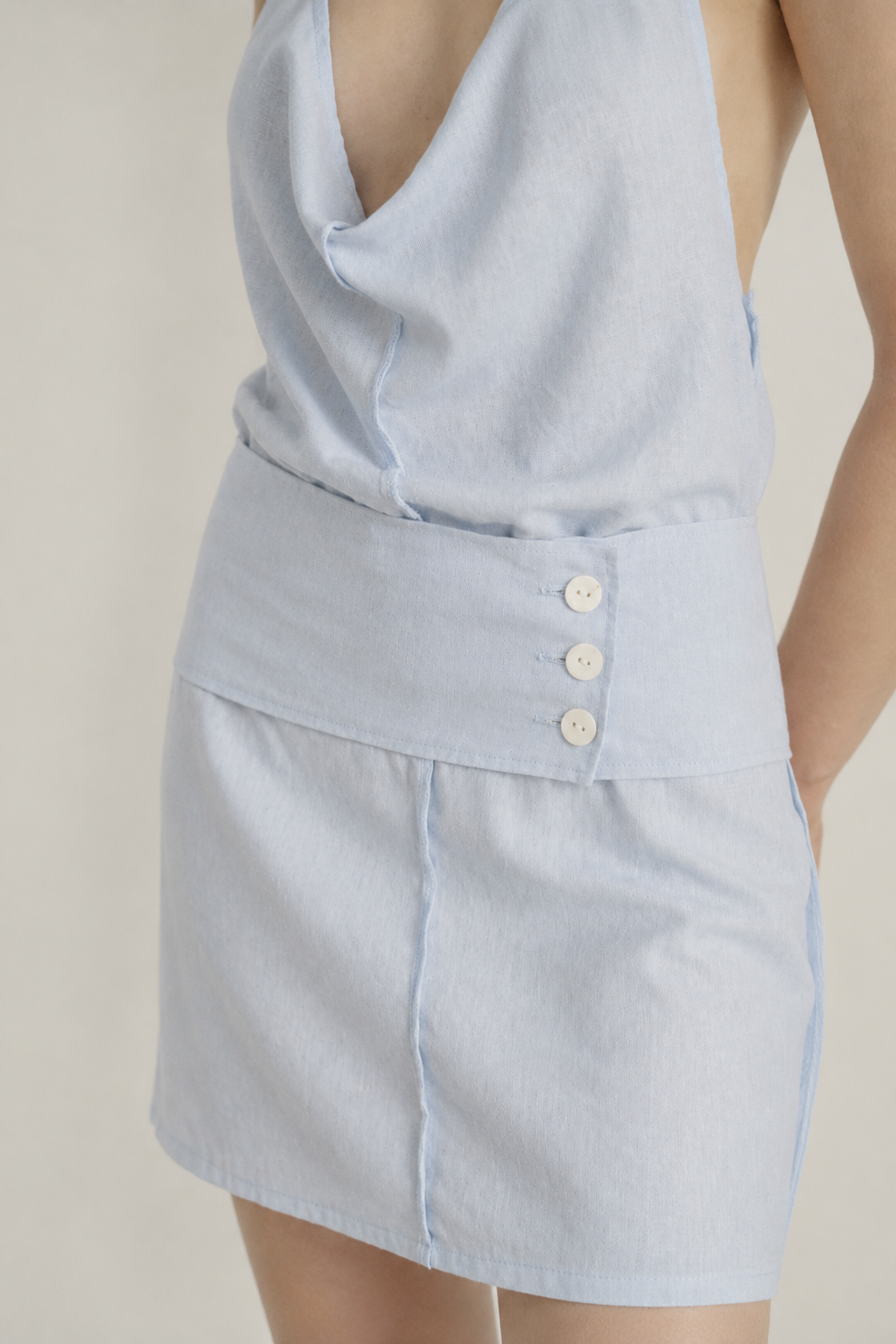 A close-up of a model wearing a light blue sleeveless top and matching high-waisted shorts with three decorative buttons on the waistband, set against a plain beige background.