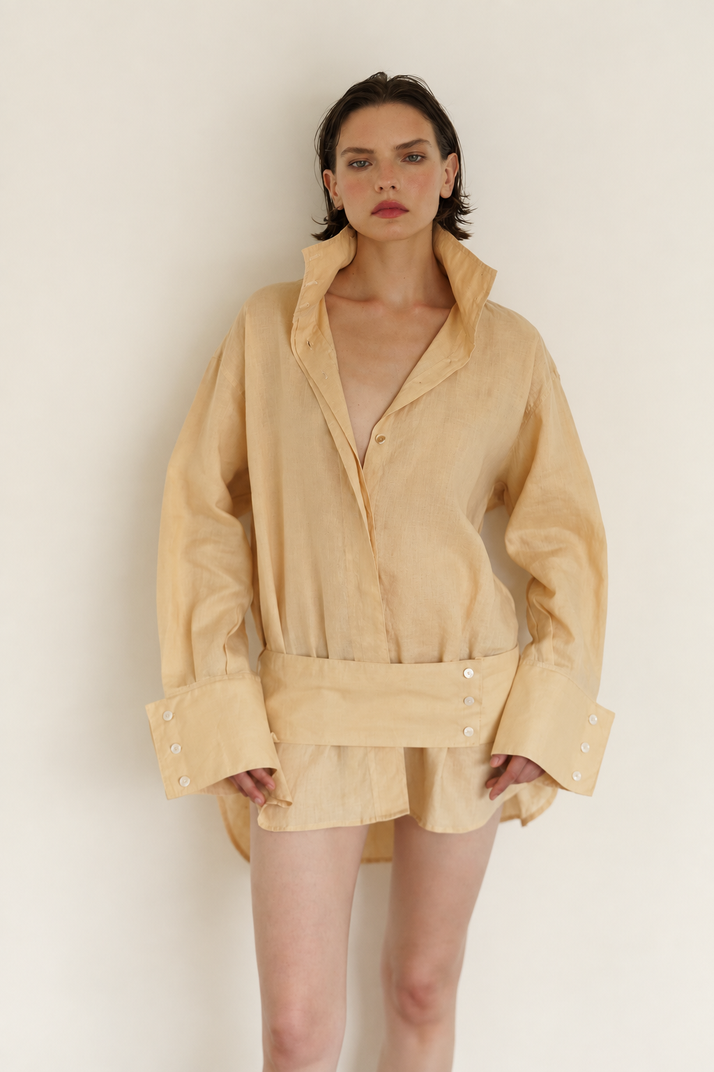 A model wearing a light beige oversized linen shirt dress with a high collar, button-down front, and a matching fabric belt with decorative buttons, set against a plain off-white background.