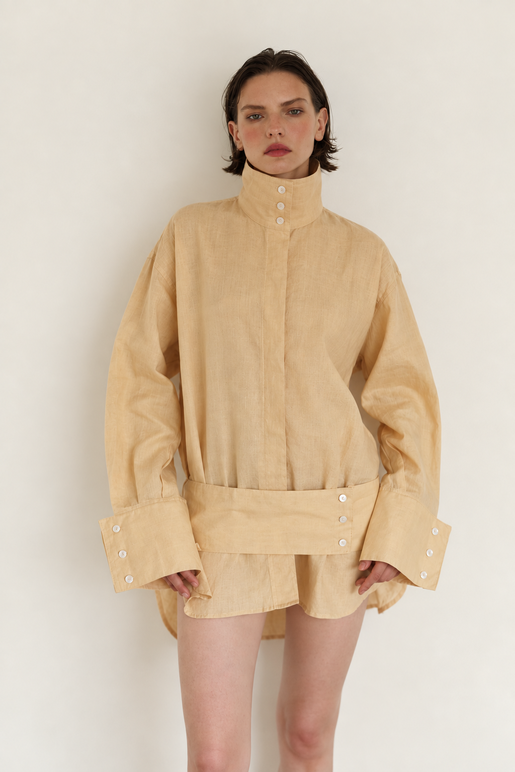 A model wearing a beige high-collar long-sleeve shirt with a matching belt and decorative buttons, set against a plain off-white background.