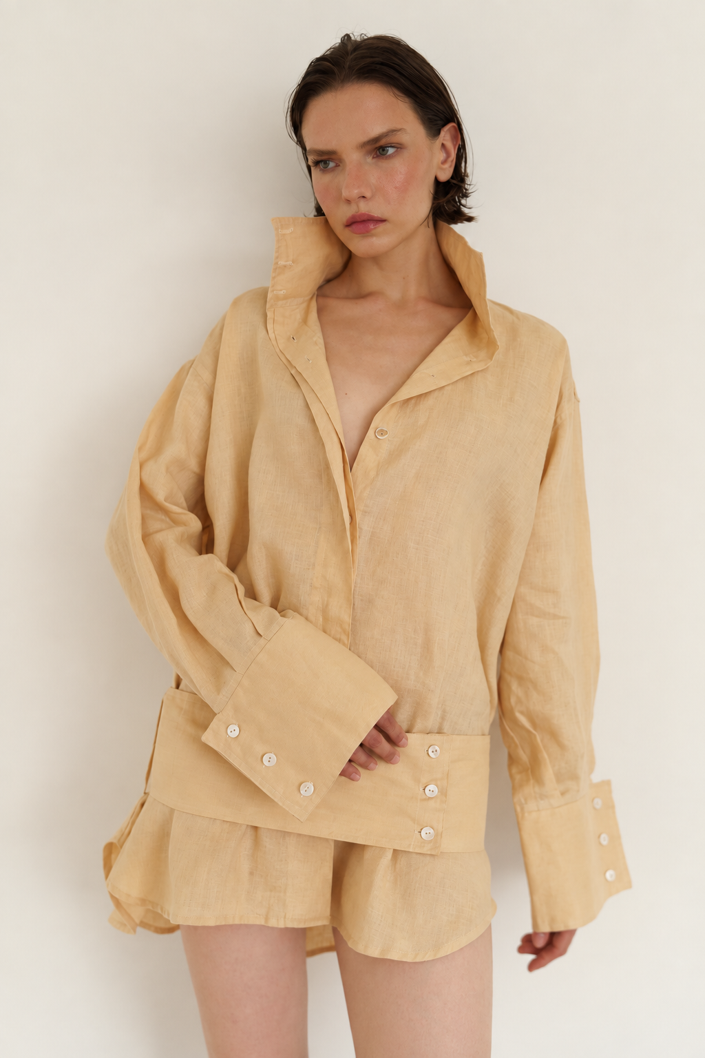 A model wearing a light beige linen shirt and matching shorts set, featuring a high collar, buttoned cuffs, and decorative white buttons, shot against a plain off-white background.