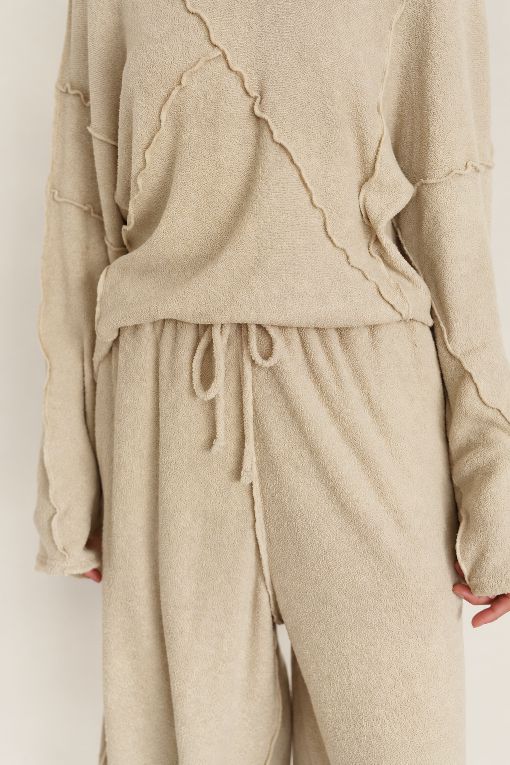 A close-up of a person wearing a matching beige knit lounge set with a long-sleeve top and drawstring pants, photographed against a plain light background.