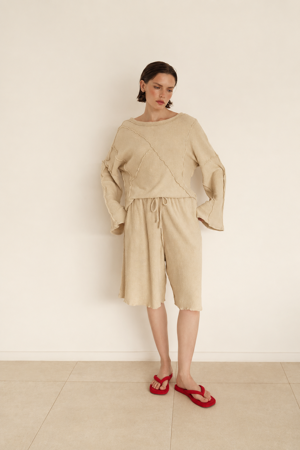 A model stands against a plain off-white wall wearing a beige two-piece linen set consisting of a long-sleeve cropped top with diagonal seam details and matching drawstring shorts, paired with red flip-flop sandals.