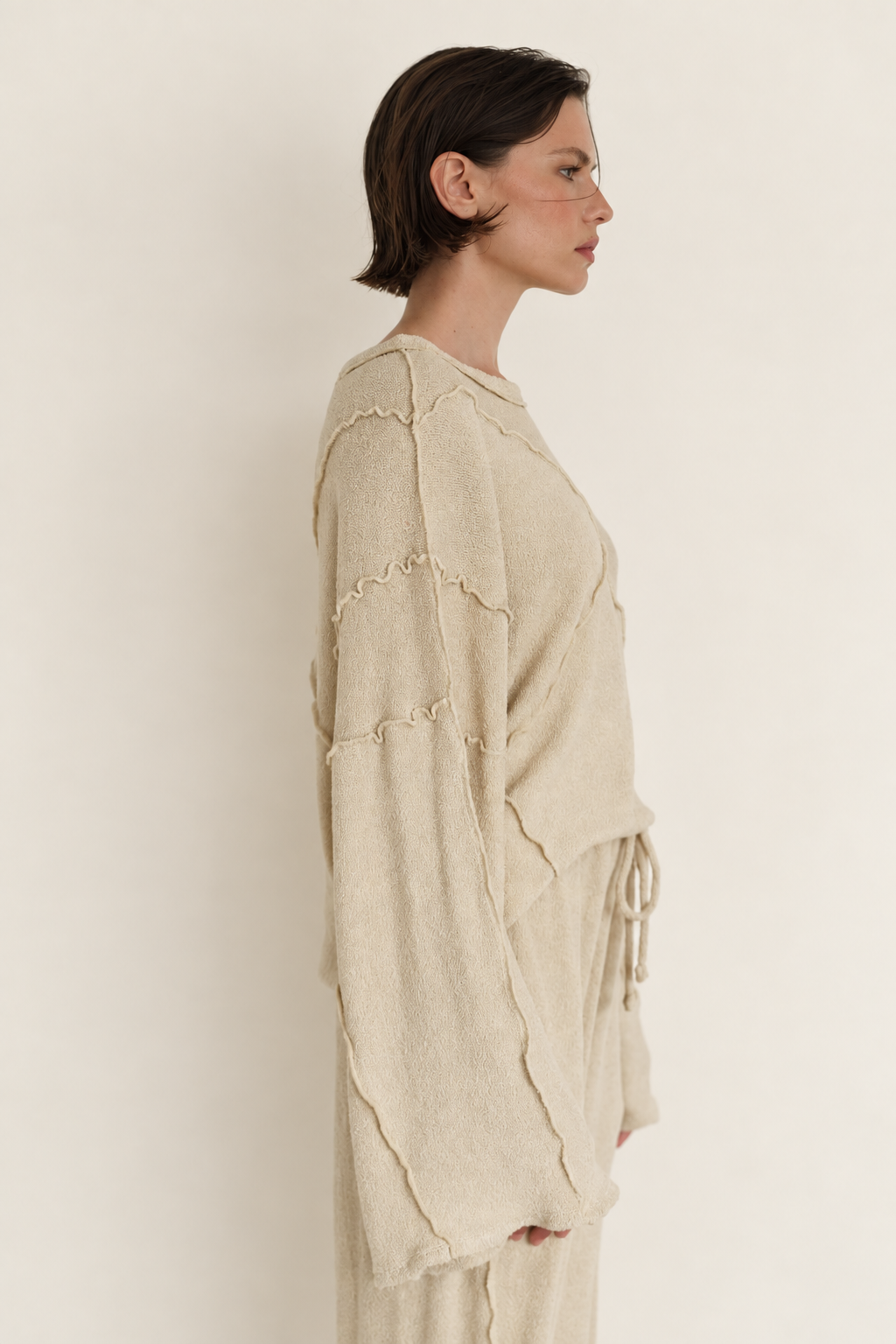 A woman shown in side profile wearing a loose, oversized cream knit top with scalloped edges and a matching drawstring tie, against a plain off-white studio background.