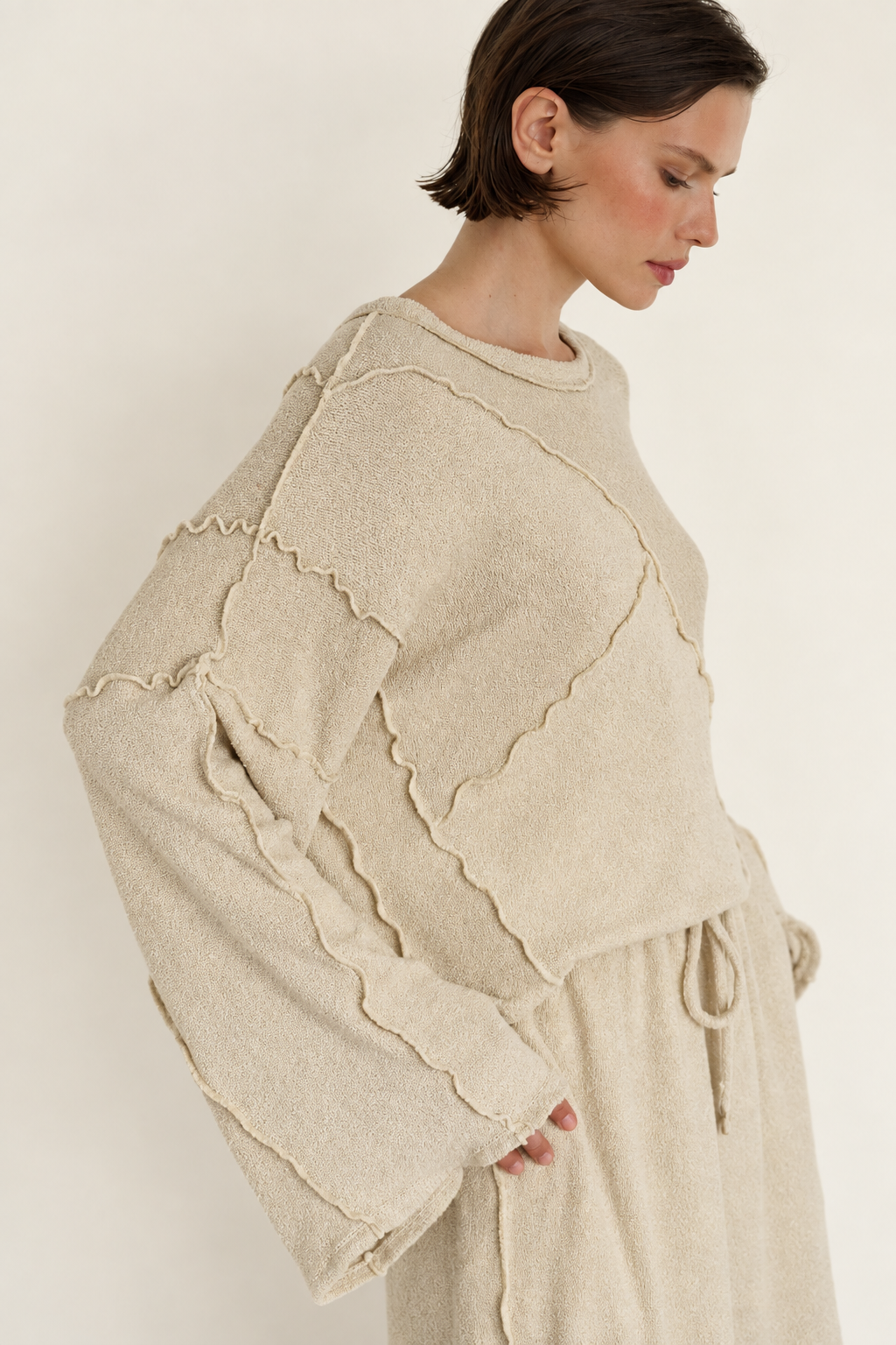 A woman wearing a cream-colored textured knit sweater with scalloped edges and a matching drawstring skirt, photographed against a plain light beige background.