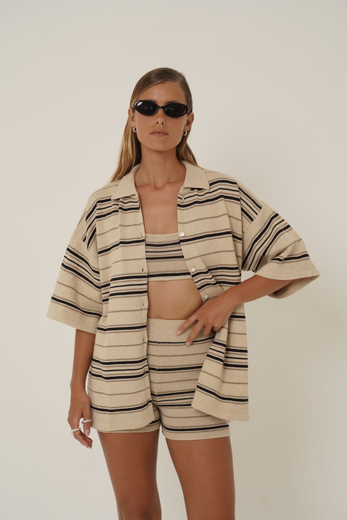 A model wearing a matching beige and black striped short-sleeve crop top and shorts set, styled with black sunglasses, against a plain light background.