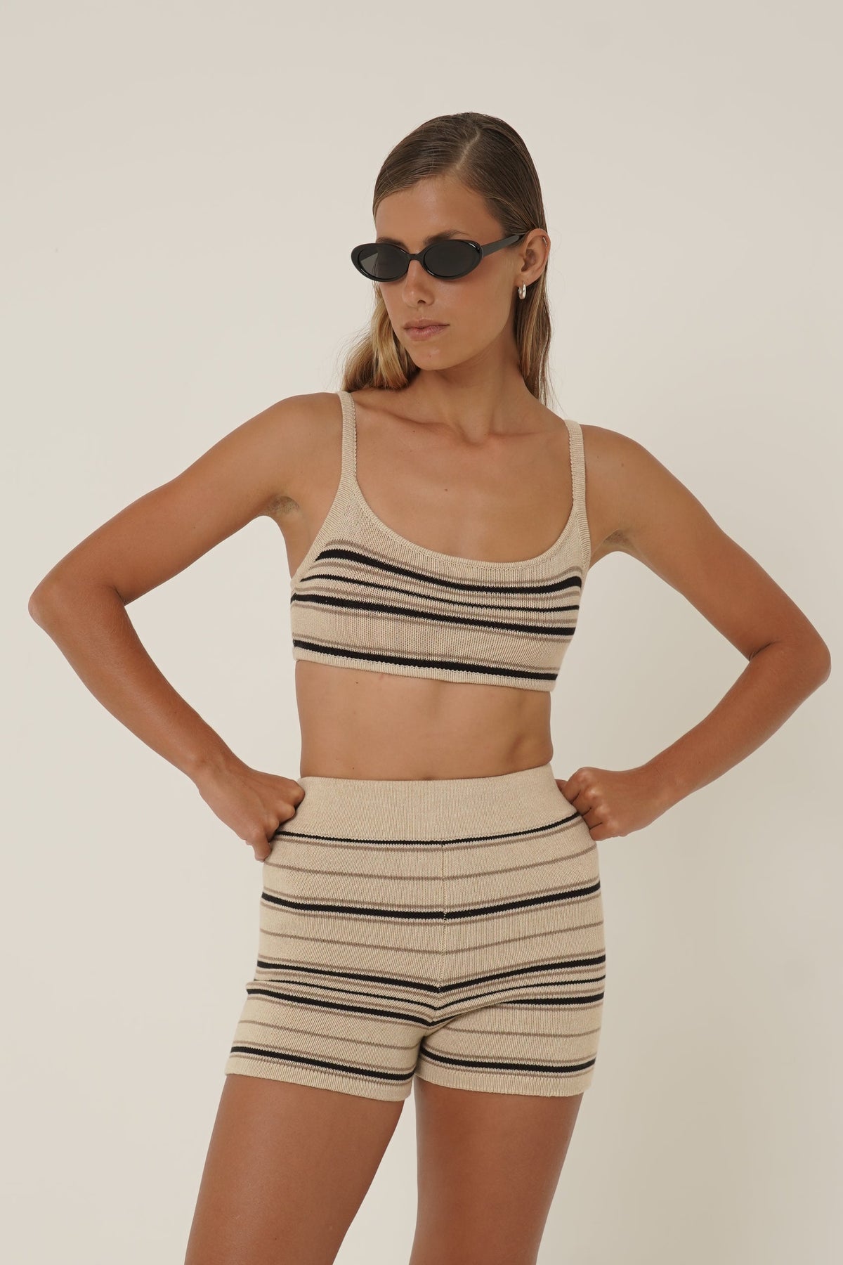 A model wearing a matching beige and black striped knit two-piece set consisting of a crop top and shorts, styled with black sunglasses, posed against a solid off-white background.
