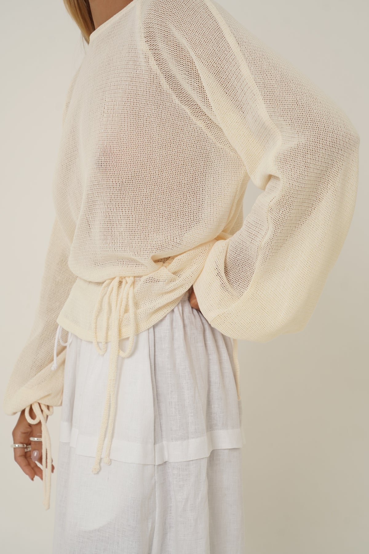 A close-up of a person wearing a loose, semi-sheer cream knit top with long balloon sleeves and a drawstring waist, paired with white wide-leg pants against a plain light beige background.