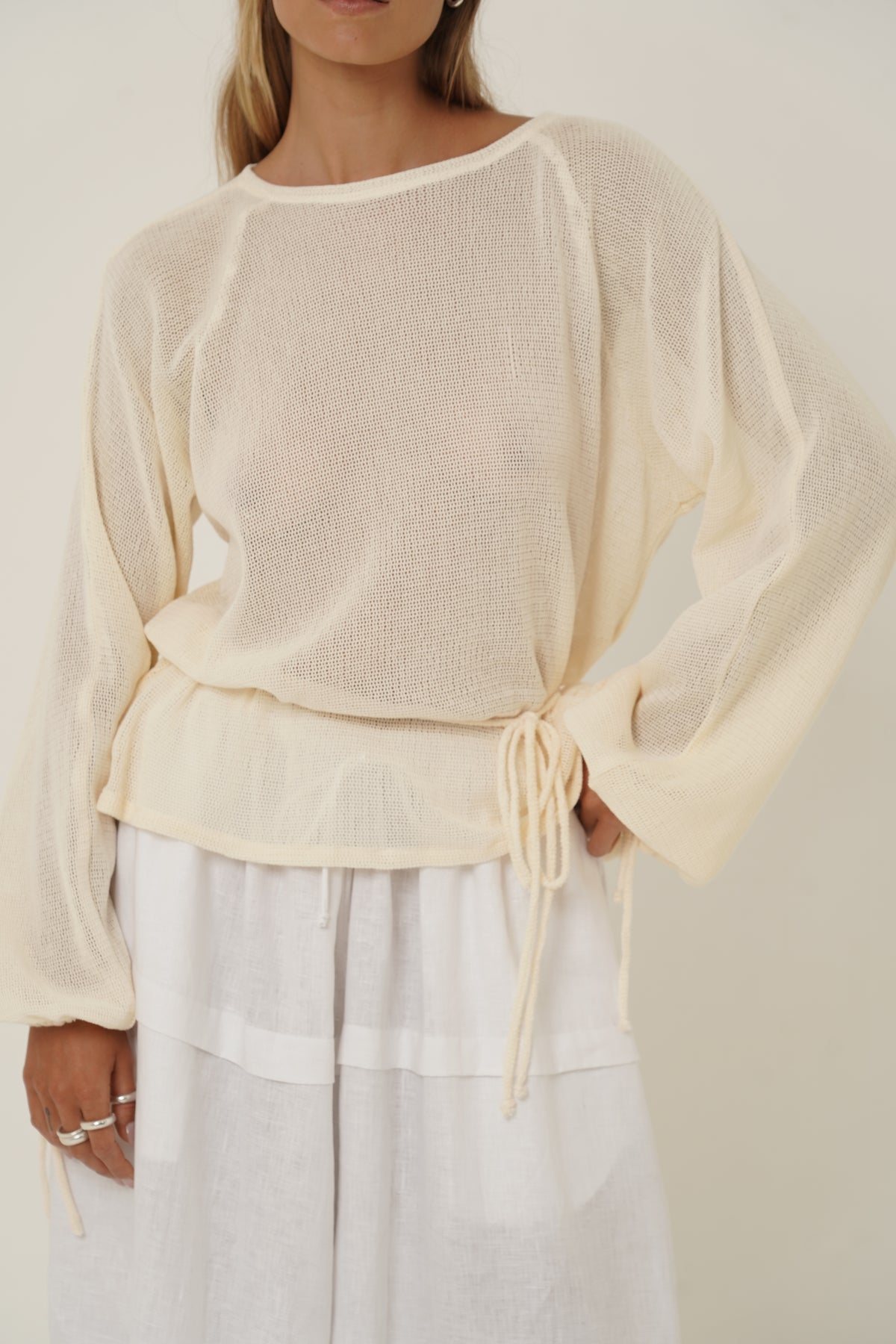 A woman wearing a cream-colored, semi-sheer long-sleeve knit top with a drawstring waist, paired with white wide-leg pants, against a plain beige background.