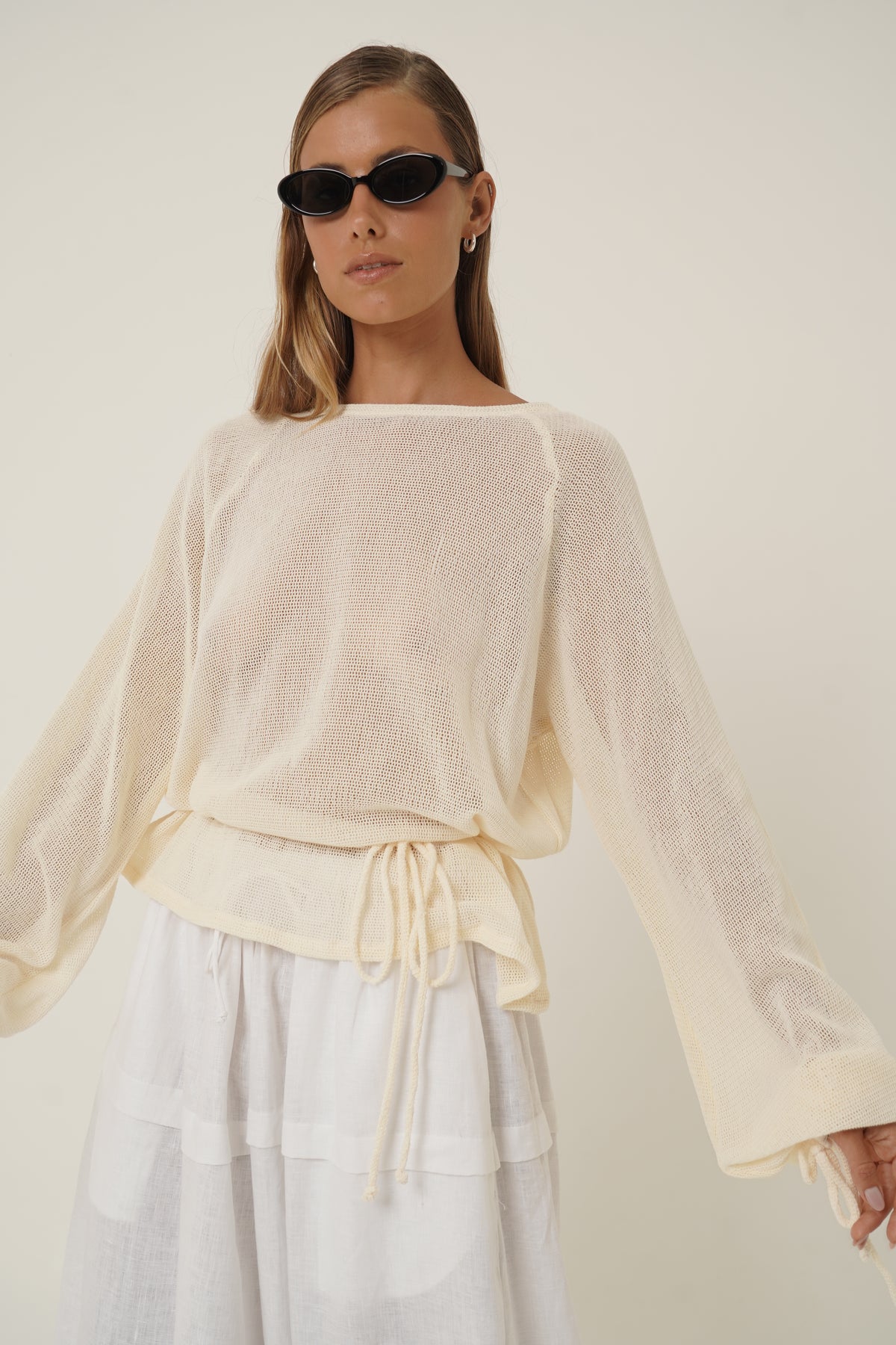 A woman modeling a cream-colored, semi-sheer long-sleeve knit top with a drawstring waist, paired with a white skirt, against a plain off-white background.