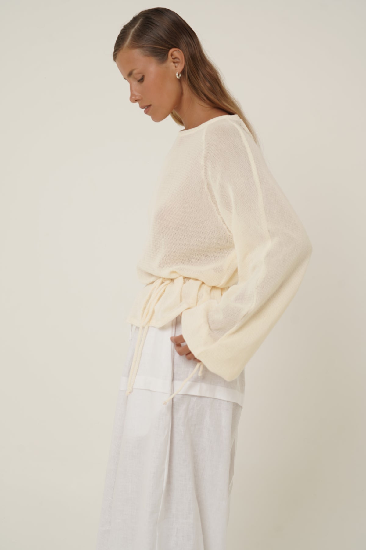 A woman wearing a cream-colored, semi-sheer long-sleeve top with a drawstring waist and white wide-leg trousers, photographed against a plain beige studio background.