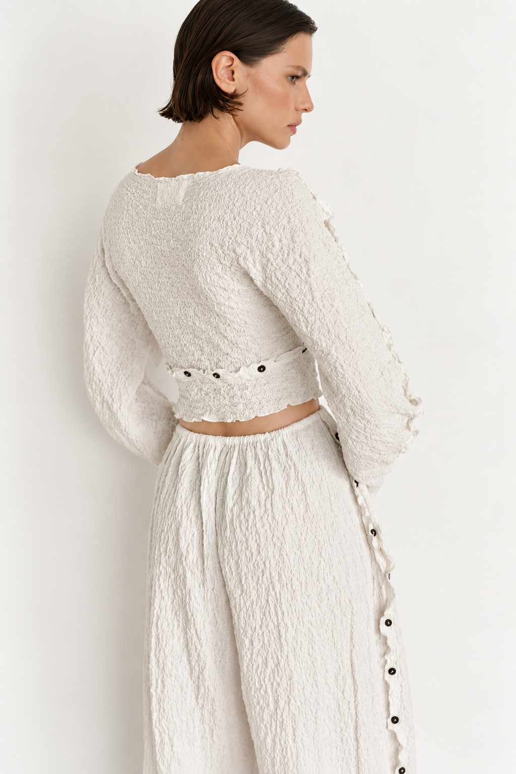 A model shown from the back wearing a textured cream two-piece outfit consisting of a cropped long-sleeve top with button details and matching high-waisted wide-leg pants with side buttons, set against a plain white background.