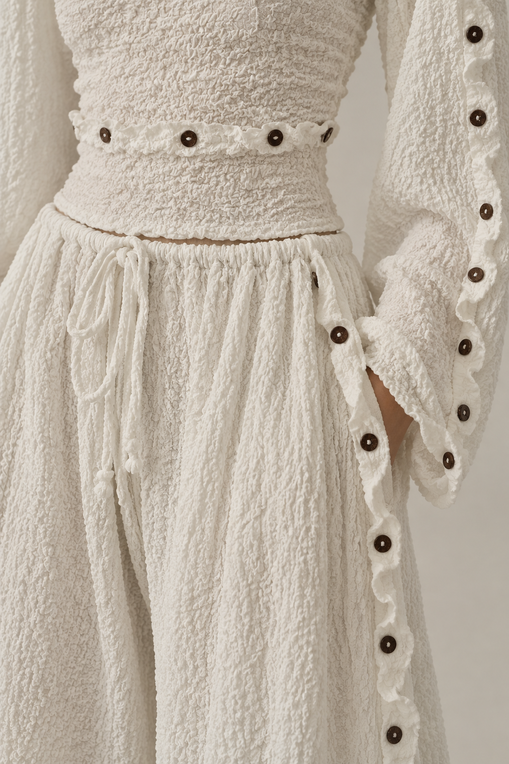 Close-up of a textured cream-colored dress featuring a matching waistband with decorative dark buttons, a drawstring waist, and long sleeves with ruffled cuffs and dark button accents.