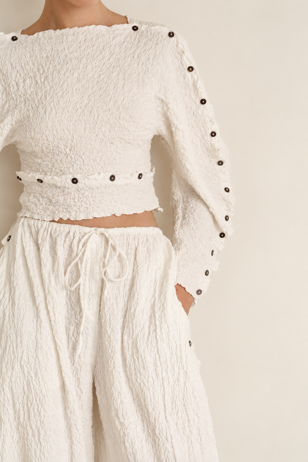 Woman wearing a white set of long pants and top with long rounded sleeves with grey button details on the seams. 