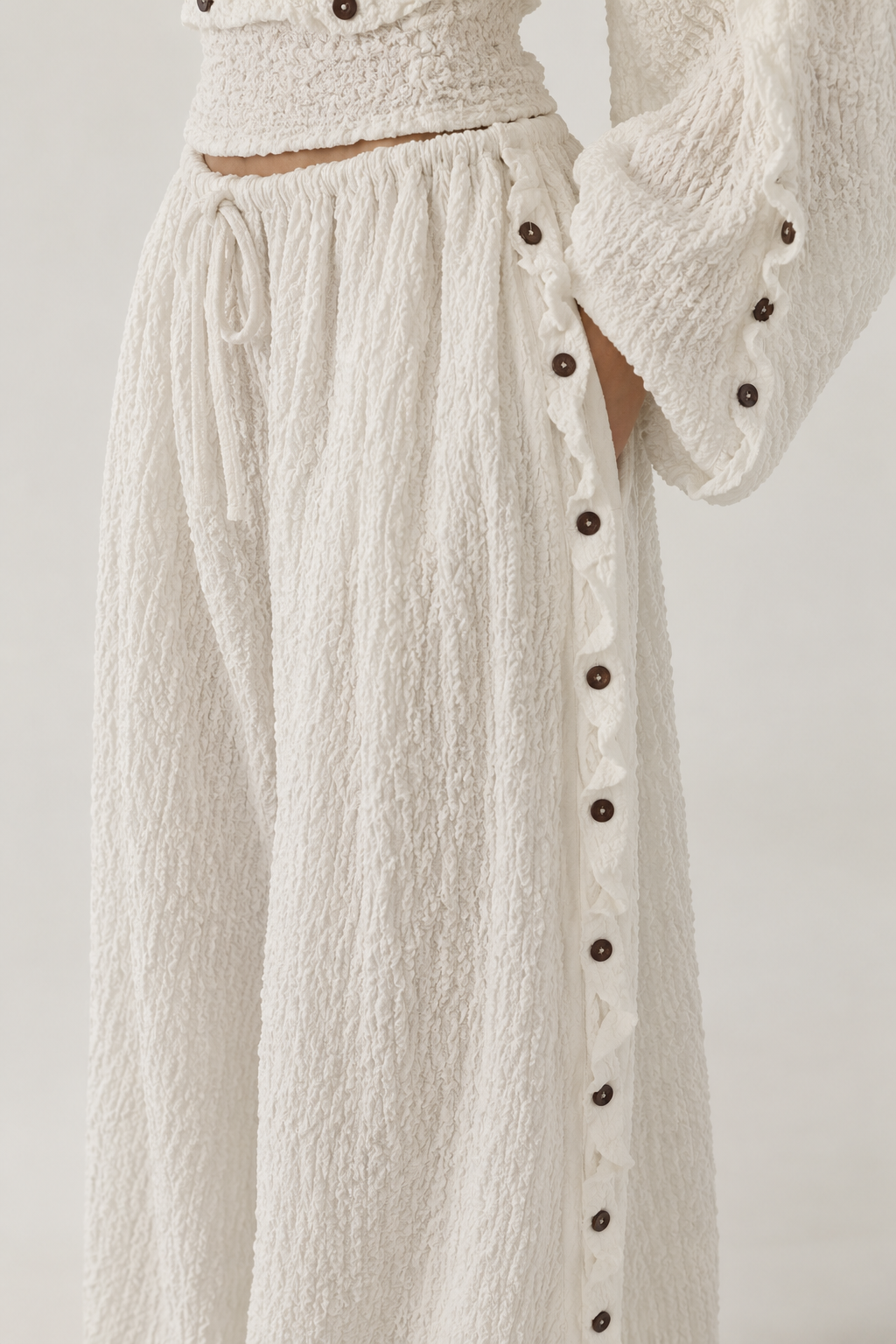 Close-up of a textured cream-colored two-piece outfit featuring wide-leg pants with a side button closure and a matching cropped top with a drawstring waist.