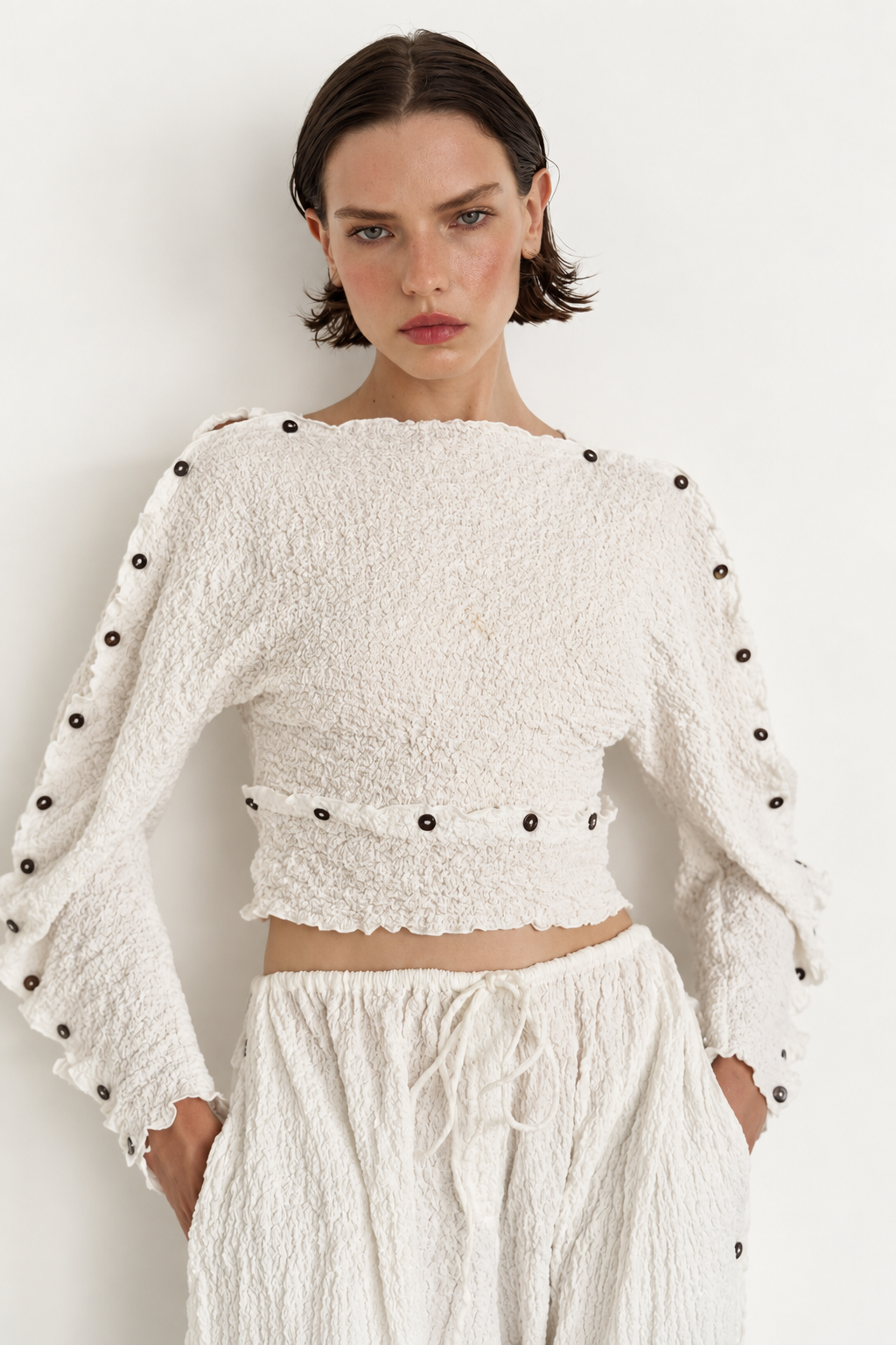 A model wearing a textured off-white cropped long-sleeve top with dark button accents down the sleeves and waist, paired with matching textured off-white pants against a plain white background.