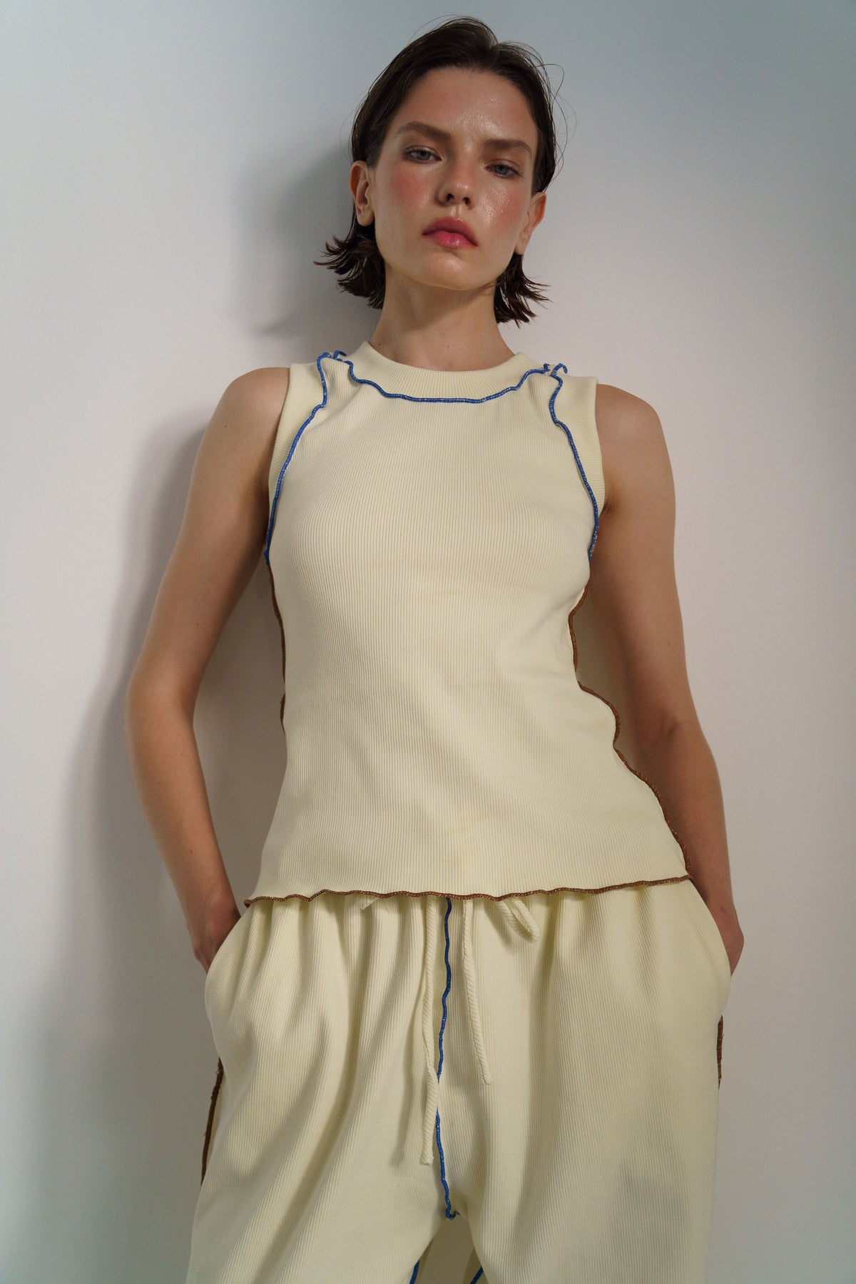 A model wearing a cream-colored ribbed knit sleeveless top and matching drawstring pants, accented with thin blue piping, set against a plain light gray wall.