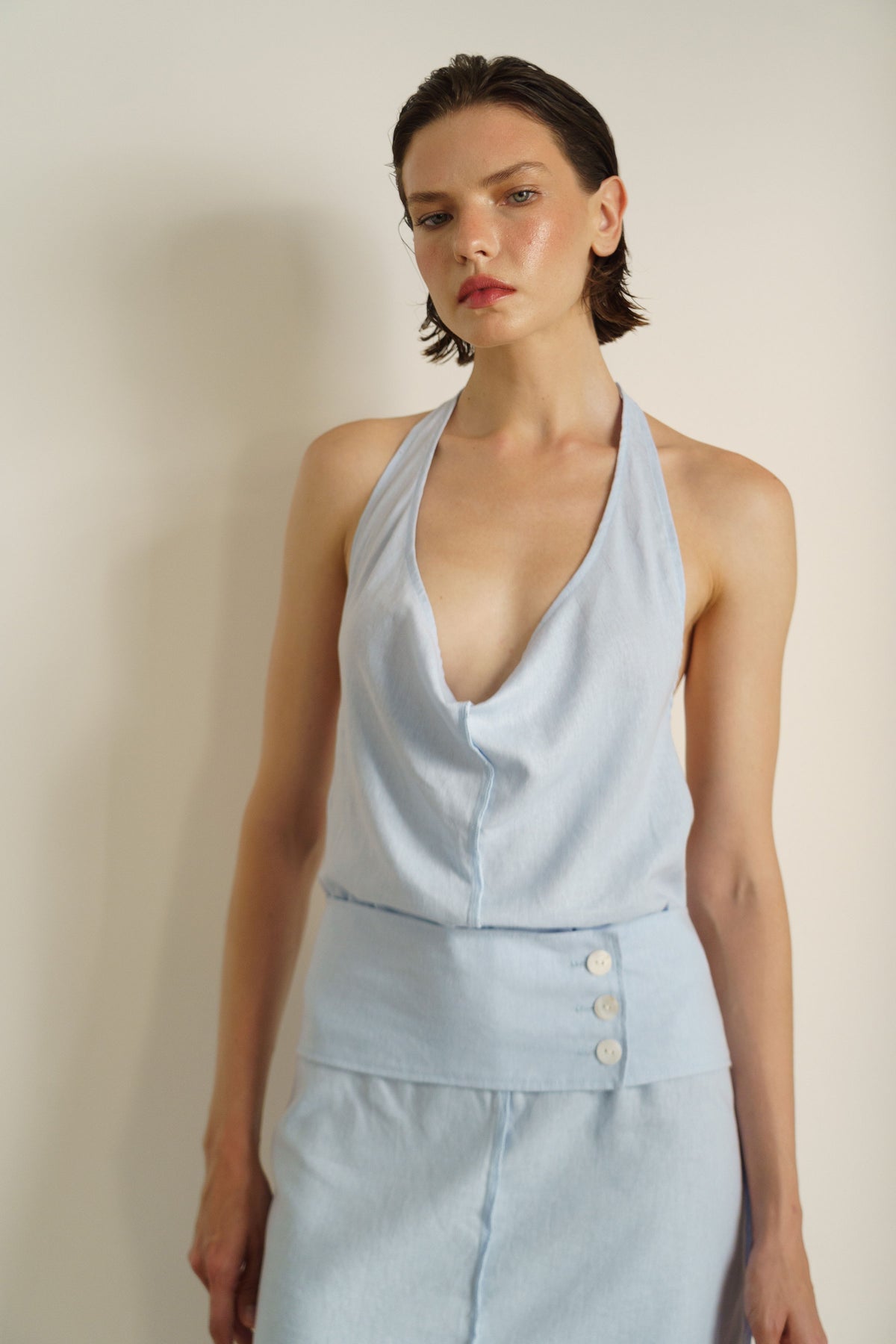 A model wearing a light blue sleeveless halter top and matching high-waisted skirt with three front buttons, set against a plain beige wall.