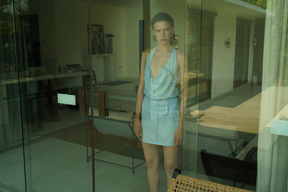 A model wearing a light blue sleeveless wrap-style top and matching mini skirt stands in front of a glass door, with a modern interior room visible behind her.