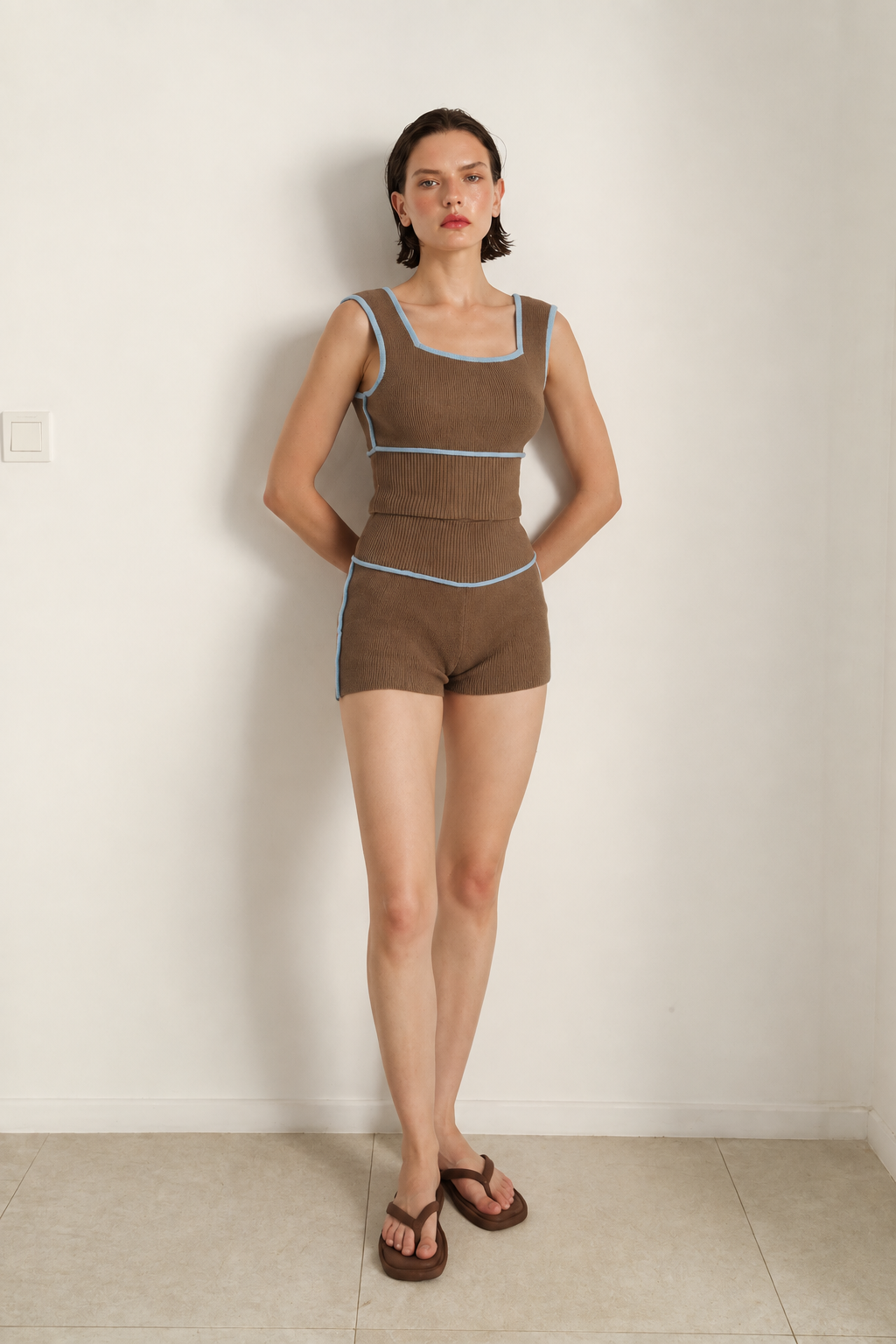 A woman stands against a plain off-white wall wearing a brown sleeveless romper with light blue contrast trim and matching brown sandals.