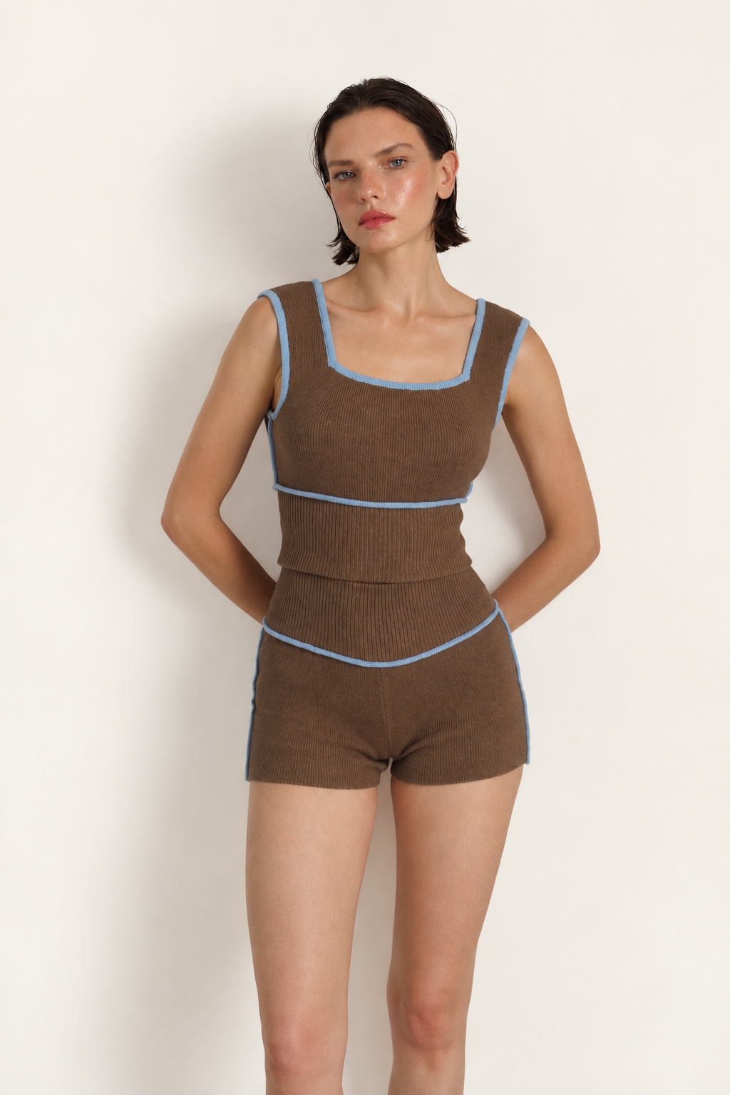 A model wearing a brown ribbed knit two-piece set consisting of a sleeveless top and matching shorts, accented with light blue piping details, set against a plain off-white background.