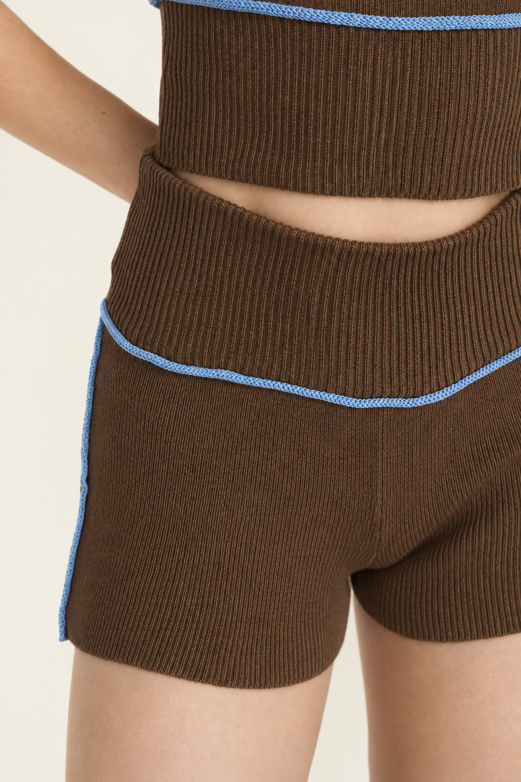 Close-up of a model wearing ribbed brown knit shorts with light blue piping trim, paired with a matching ribbed top against a plain light background.
