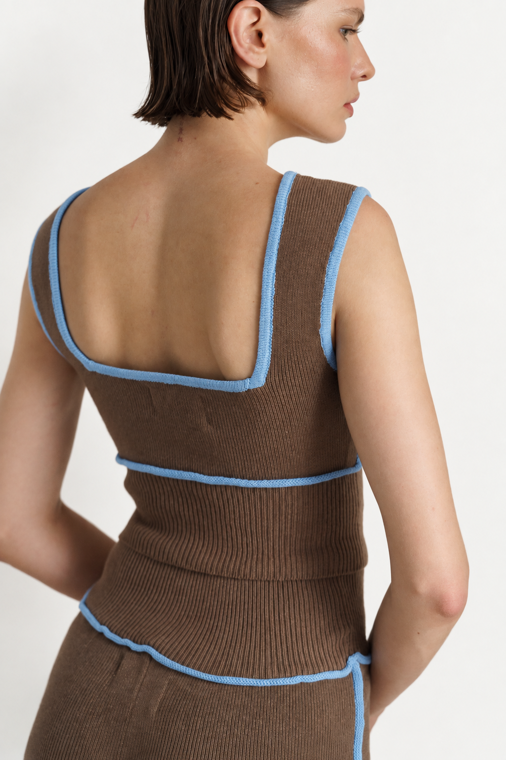 A woman shown from the back wearing a brown ribbed knit sleeveless top with light blue trim, against a plain light background.
