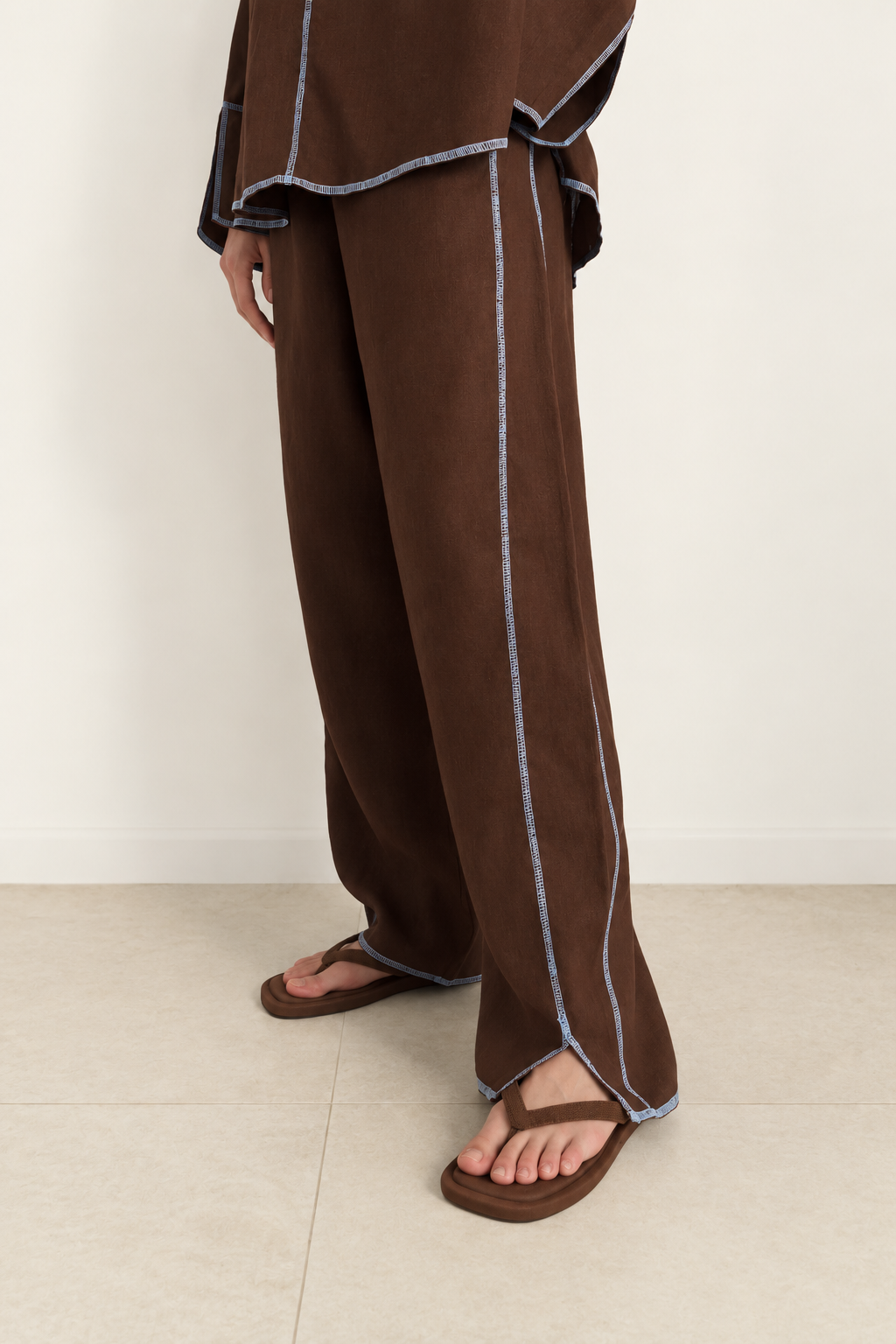 A person wearing loose brown wide-leg pants with contrasting light blue-gray piping, paired with brown sandals, standing against a plain off-white wall and light beige floor.