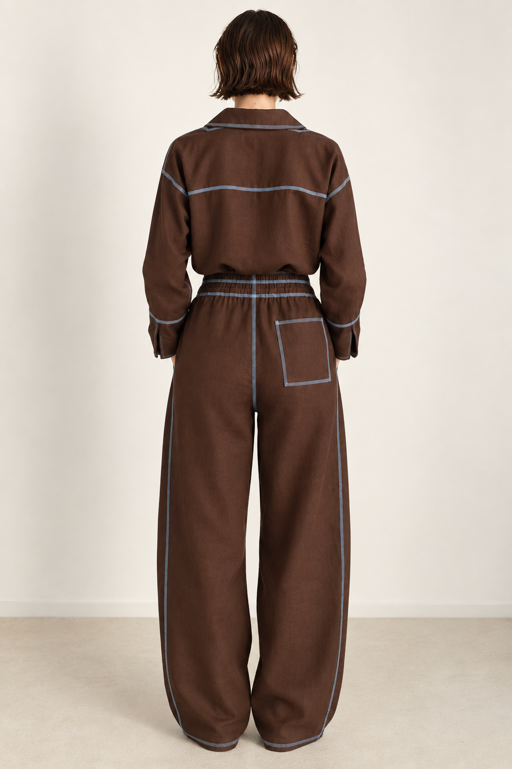 A model viewed from the back wearing a brown long-sleeve jumpsuit with light blue contrast stitching, set against a plain off-white studio background.