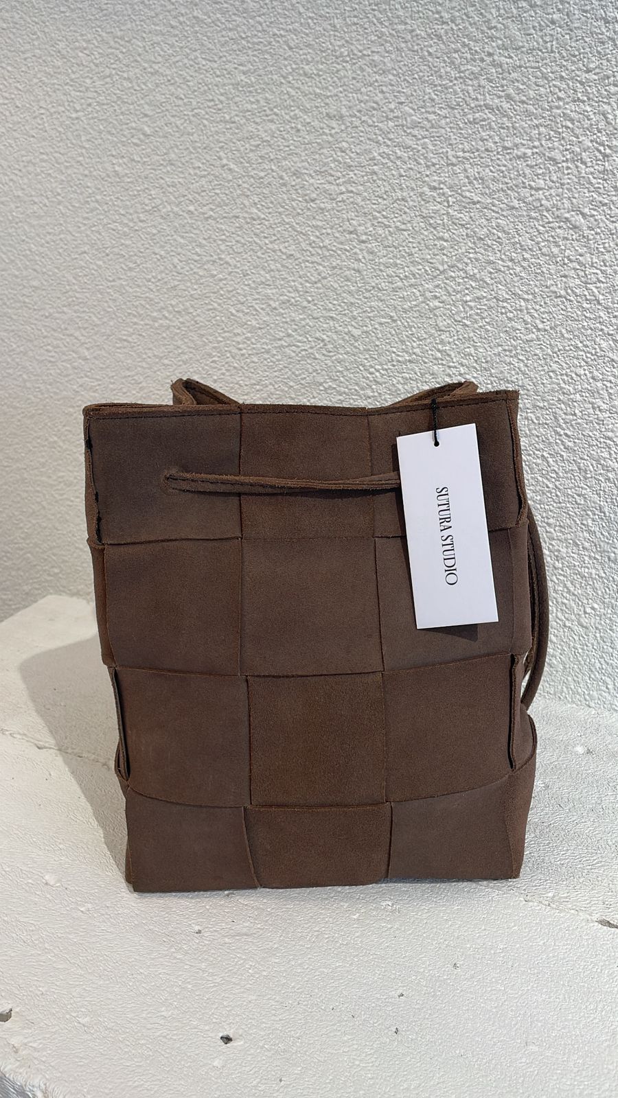 A brown woven leather bucket bag with a white paper tag that reads 'SUTURA  STUDIO', set against a textured off-white wall.