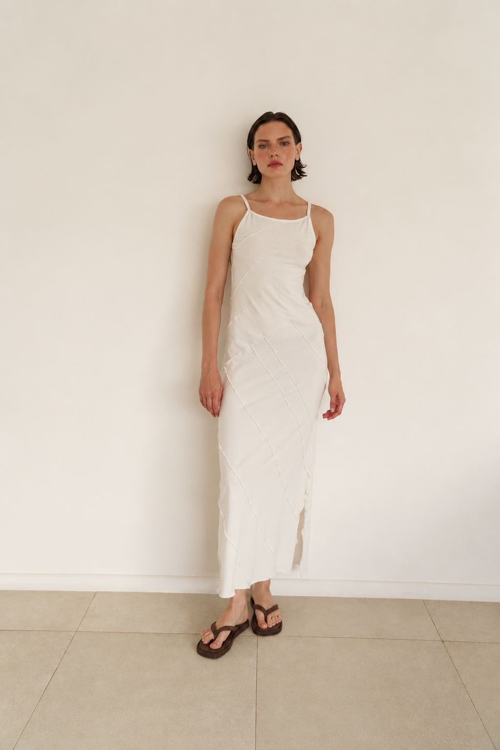 A woman wearing a white spaghetti-strap midi slip dress with a side slit, paired with dark brown sandals, standing against a plain white wall.