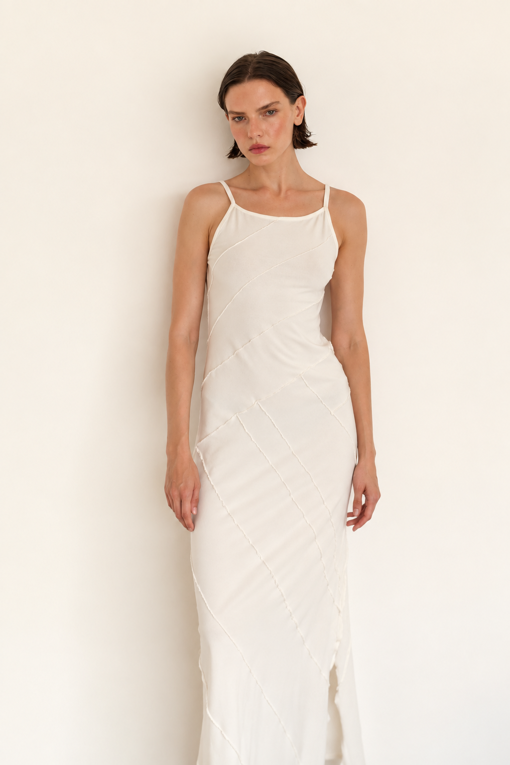 A woman modeling a white sleeveless maxi dress with thin straps and a subtle textured geometric pattern, standing against a plain off-white wall.