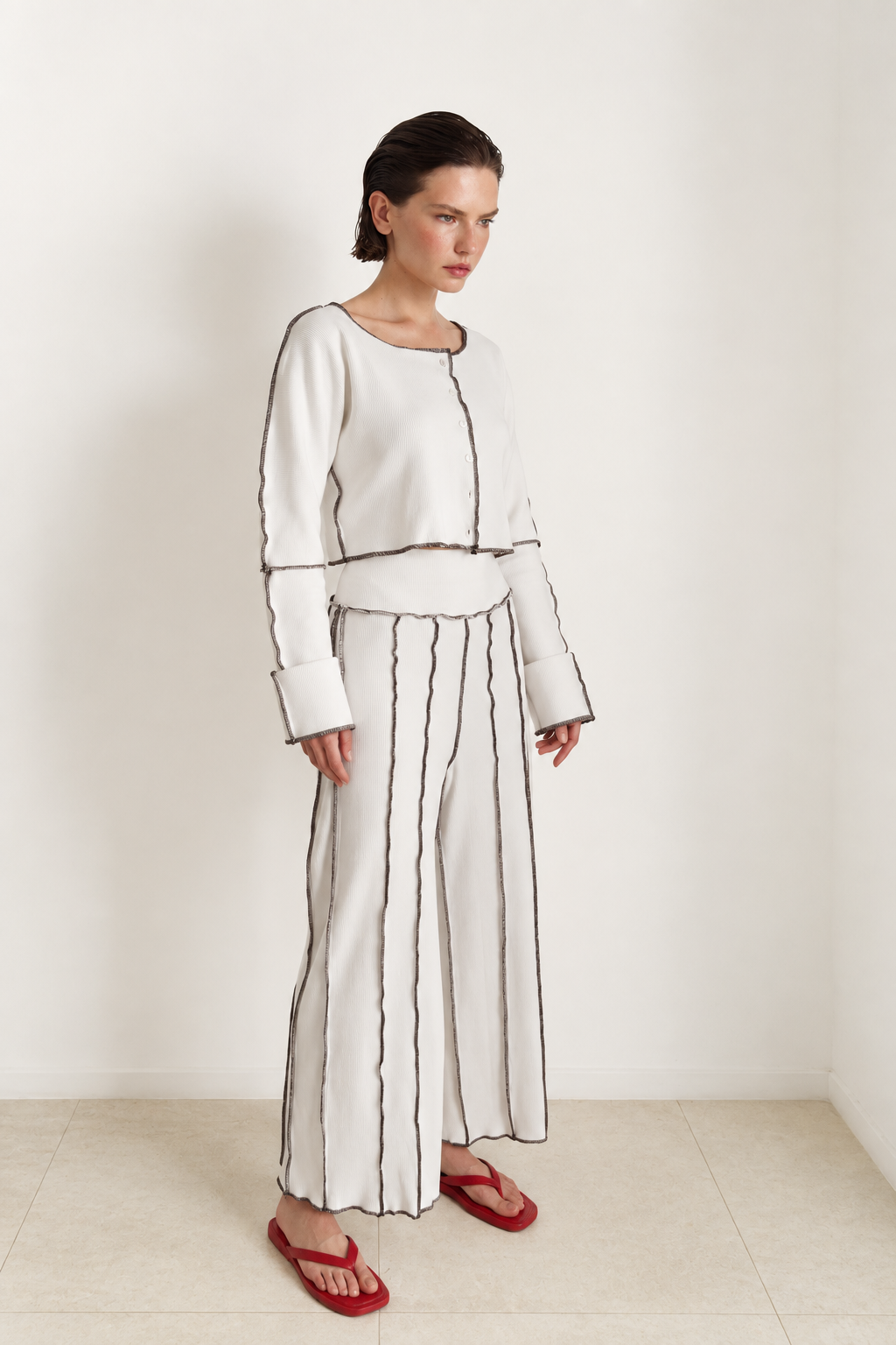 A woman modeling a white two-piece outfit consisting of a cropped long-sleeve top and matching wide-leg trousers, both detailed with thin black vertical stripes and piping, paired with red sandals.