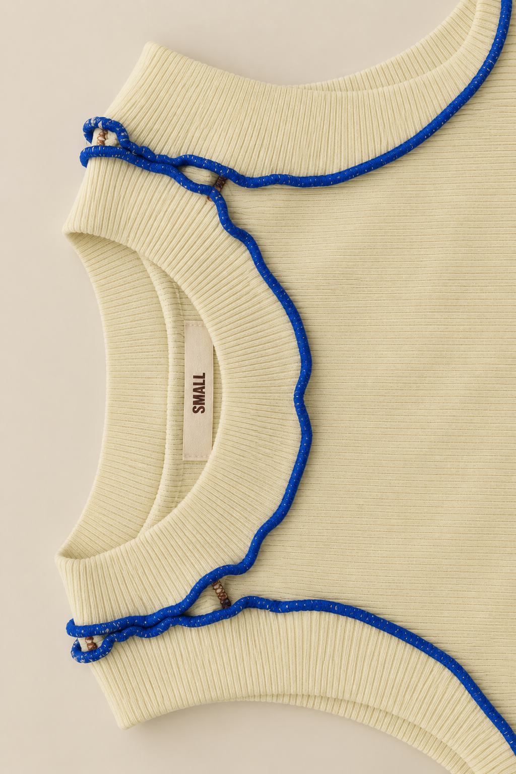 Close-up of a cream ribbed knit top with a scalloped blue cord trim detail, showing a small brand label that reads 'SMALL'.